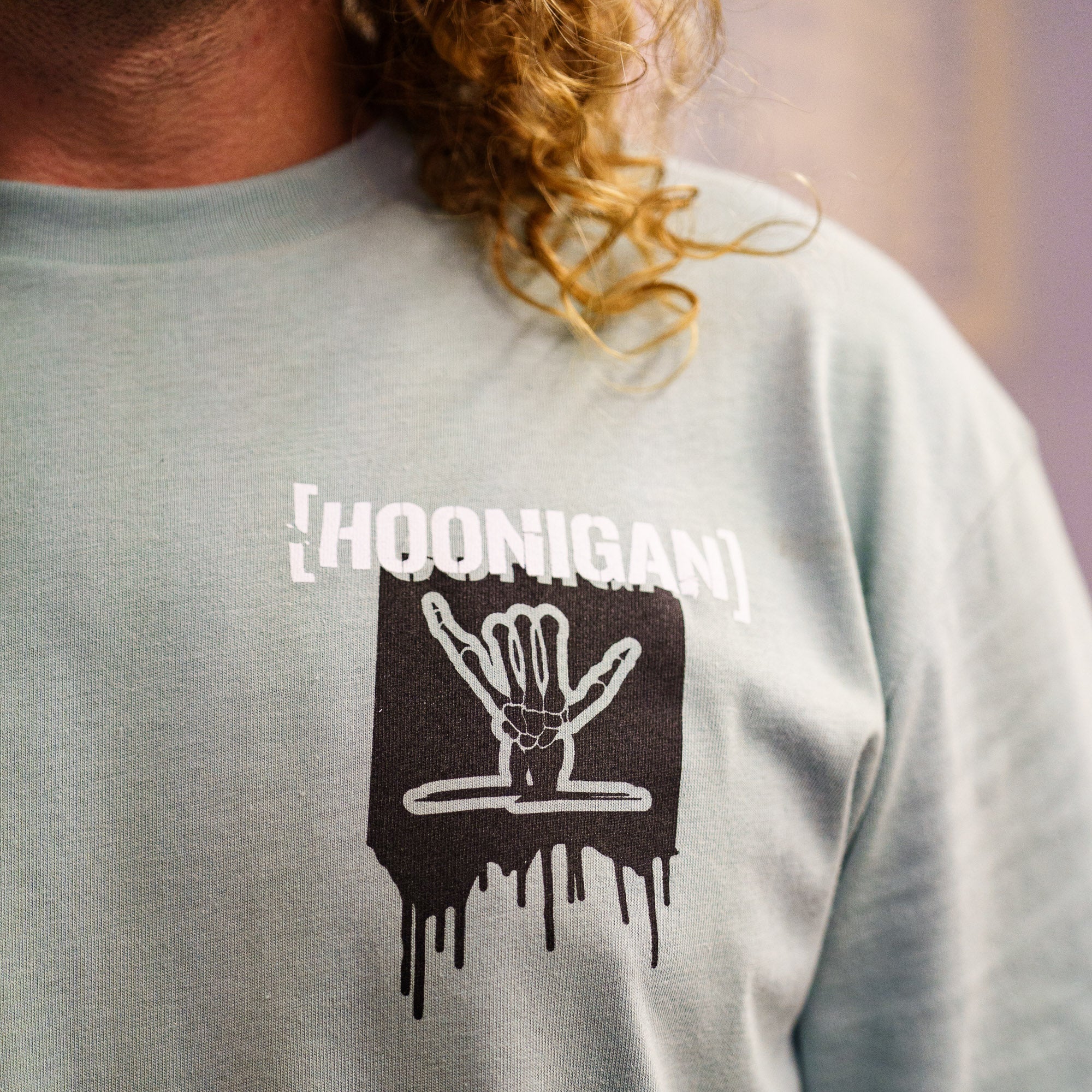 Light green short sleeve t-shirt featuring a graphic of a hand and the Hoonigan logo, perfect for motorsport enthusiasts.