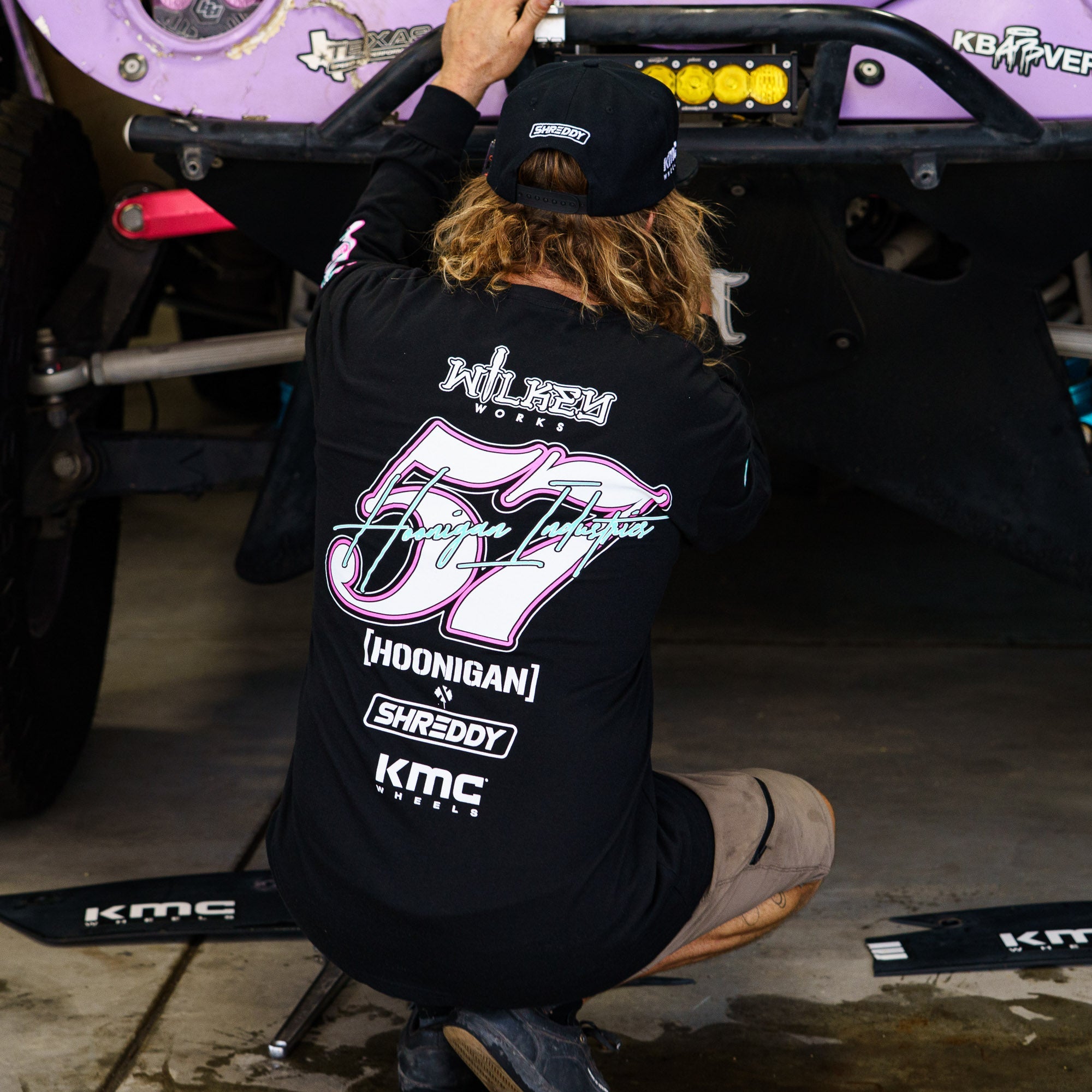 A person kneels beside a vehicle, wearing a black long sleeve Hoonigan x Shreddy 57 T-shirt, showcasing automotive passion.
