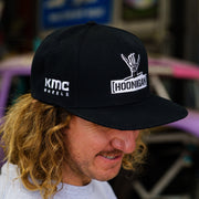 Black snapback hat featuring Hoonigan logo and KMC Wheels branding, worn by a man with curly hair.