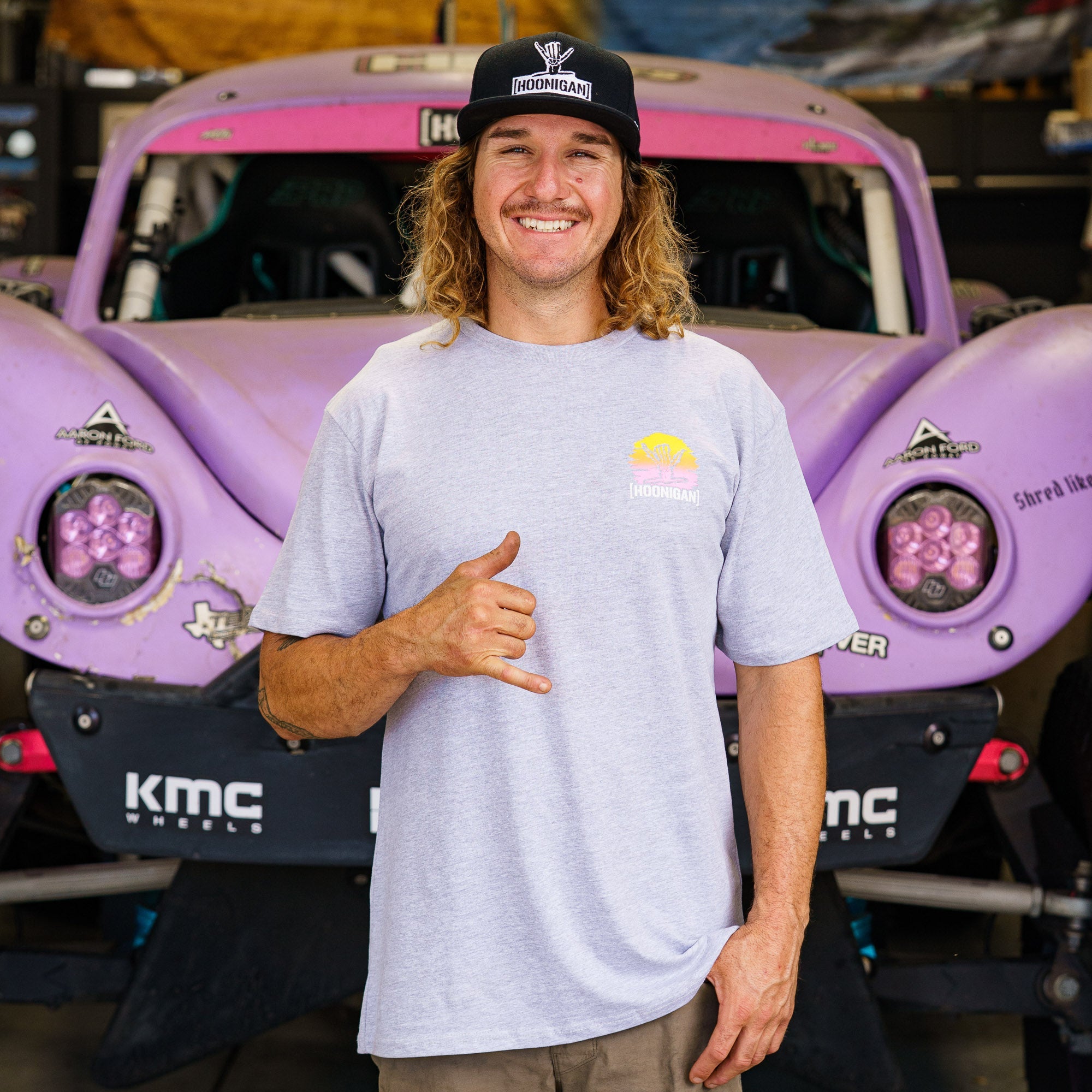 Model wearing a light gray Hoonigan x Shreddy Desert Shred short sleeve t-shirt in front of a purple off-road vehicle.