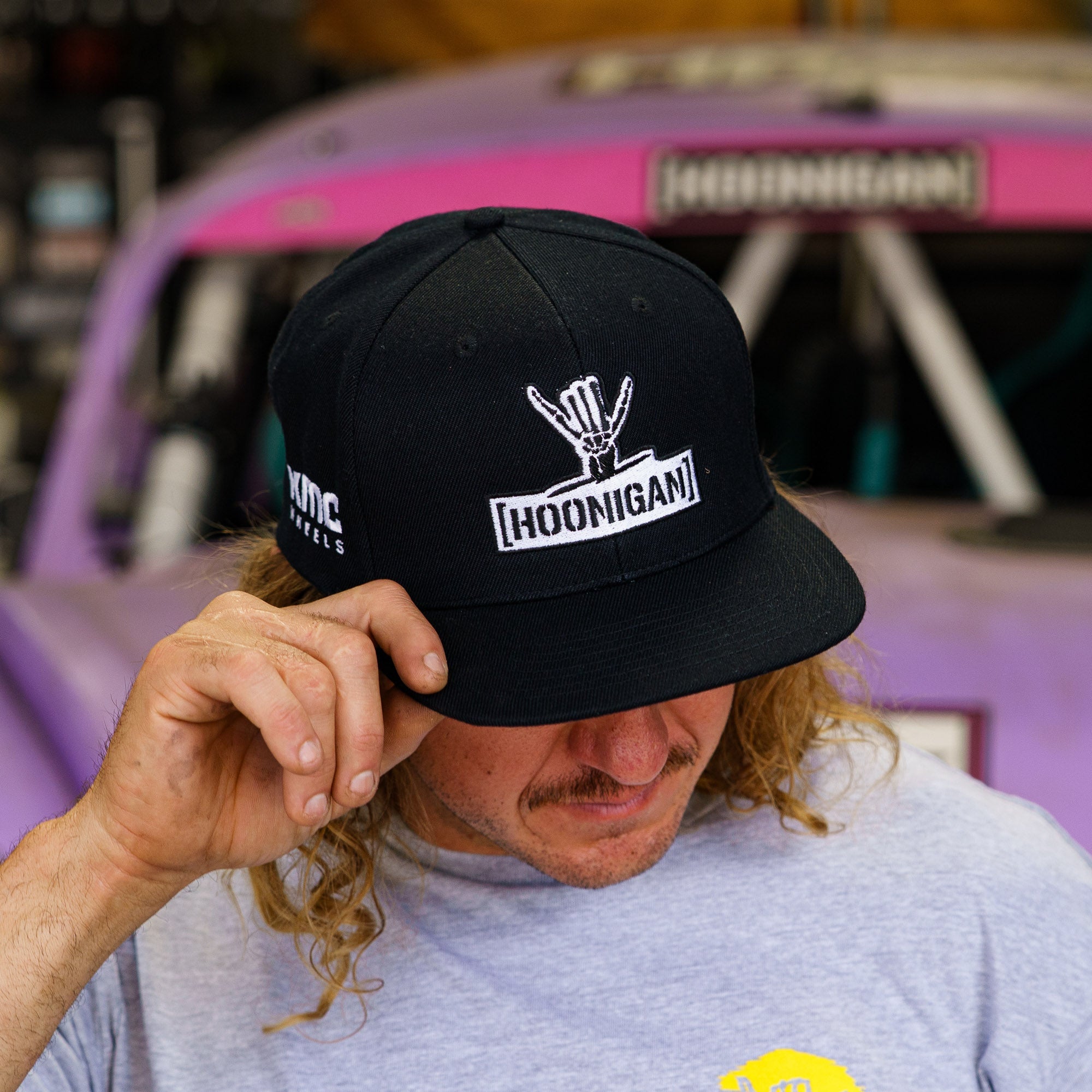Hoonigan x Shreddy Snapback Hat worn by a man, featuring a logo and a black design, set against a motorsport backdrop.