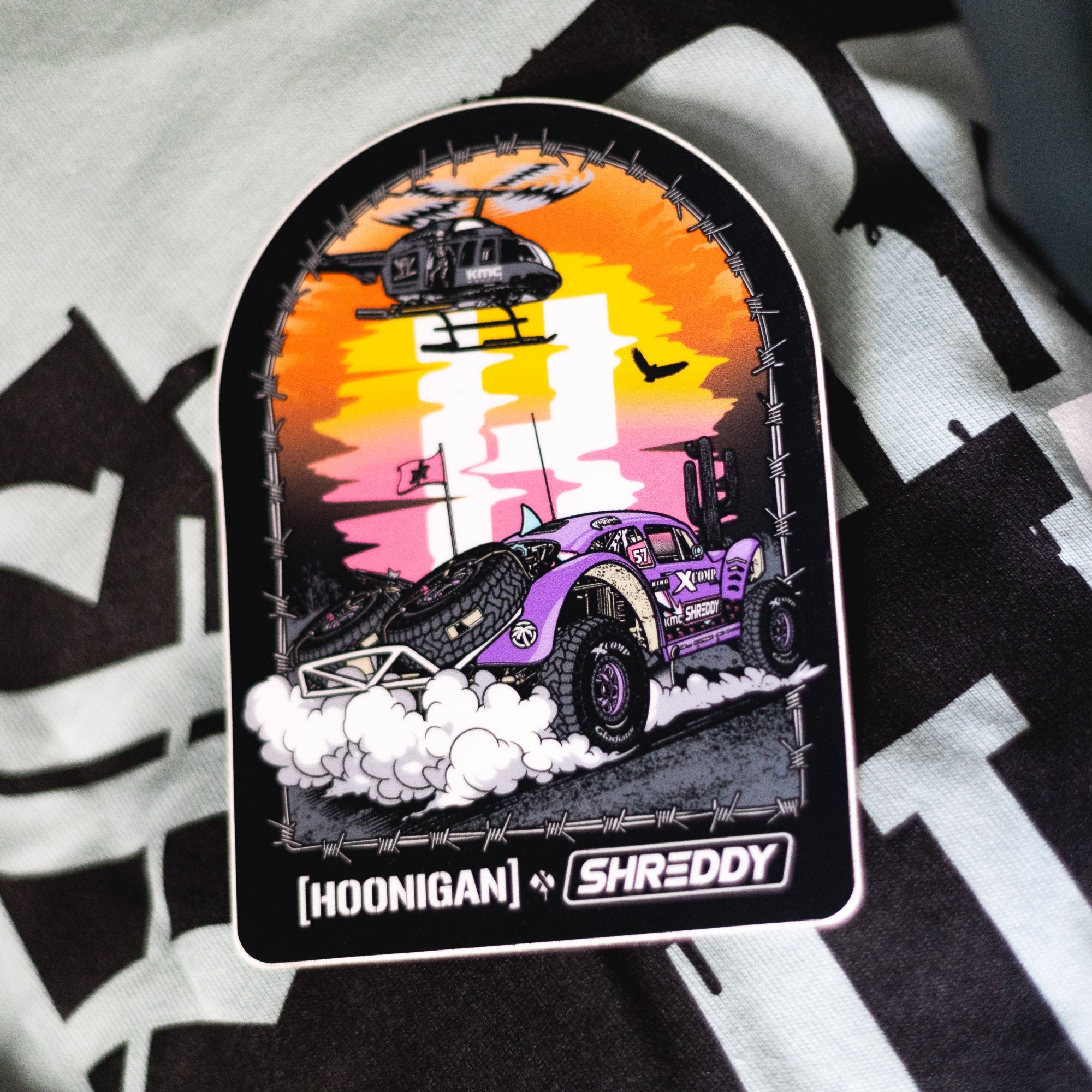 Hoonigan x Shreddy DESERT Sticker featuring a vibrant off-road vehicle and helicopter against a sunset backdrop.