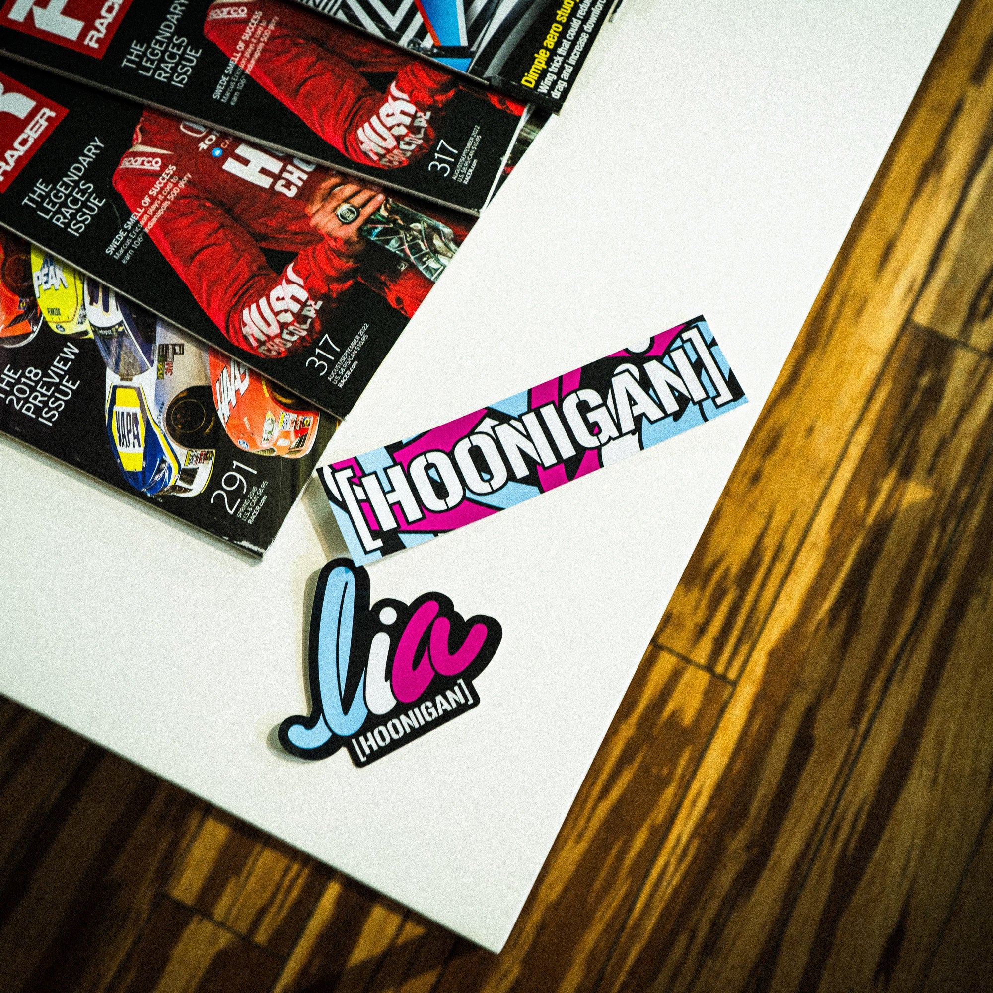 Hoonigan x Lia Block HELMET CENSOR BAR Sticker displayed on a table with automotive magazines in the background.