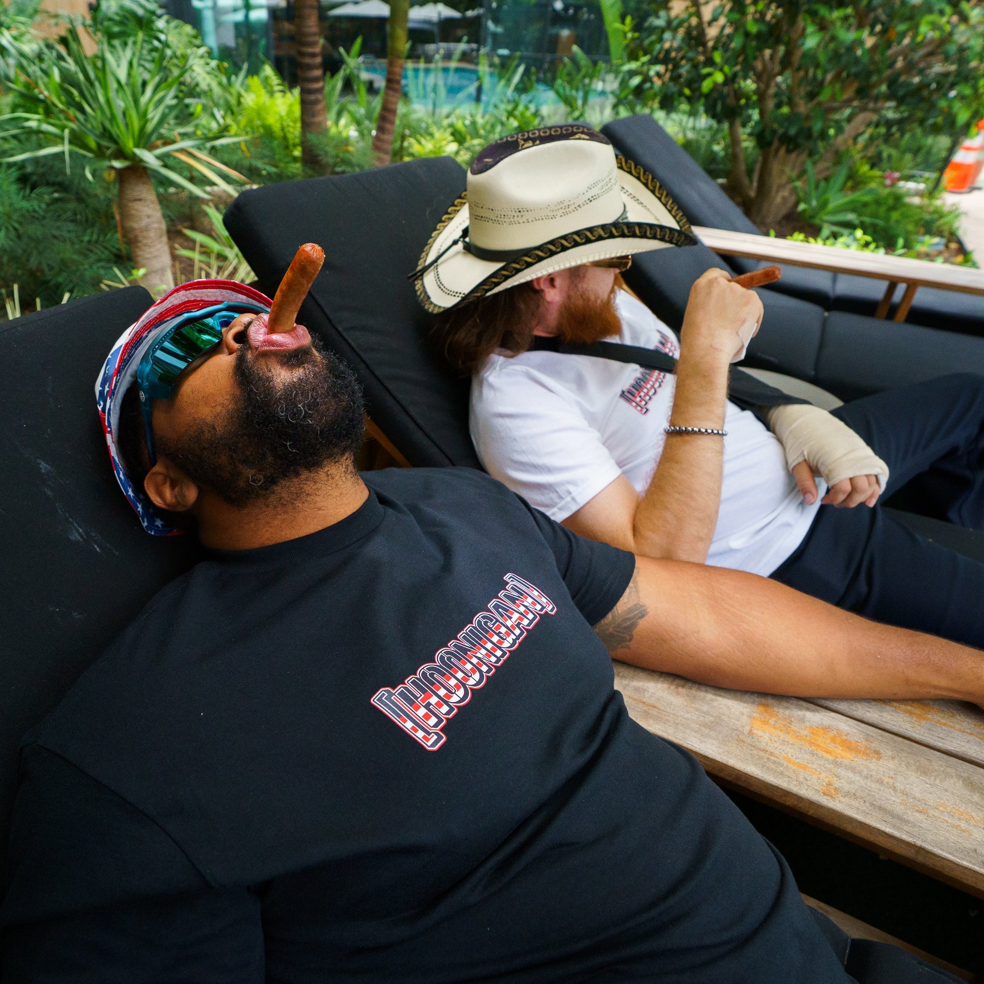 Two men relaxing on lounge chairs outdoors, one wearing a Hoonigan T-shirt and the other in a white shirt with a large hat.