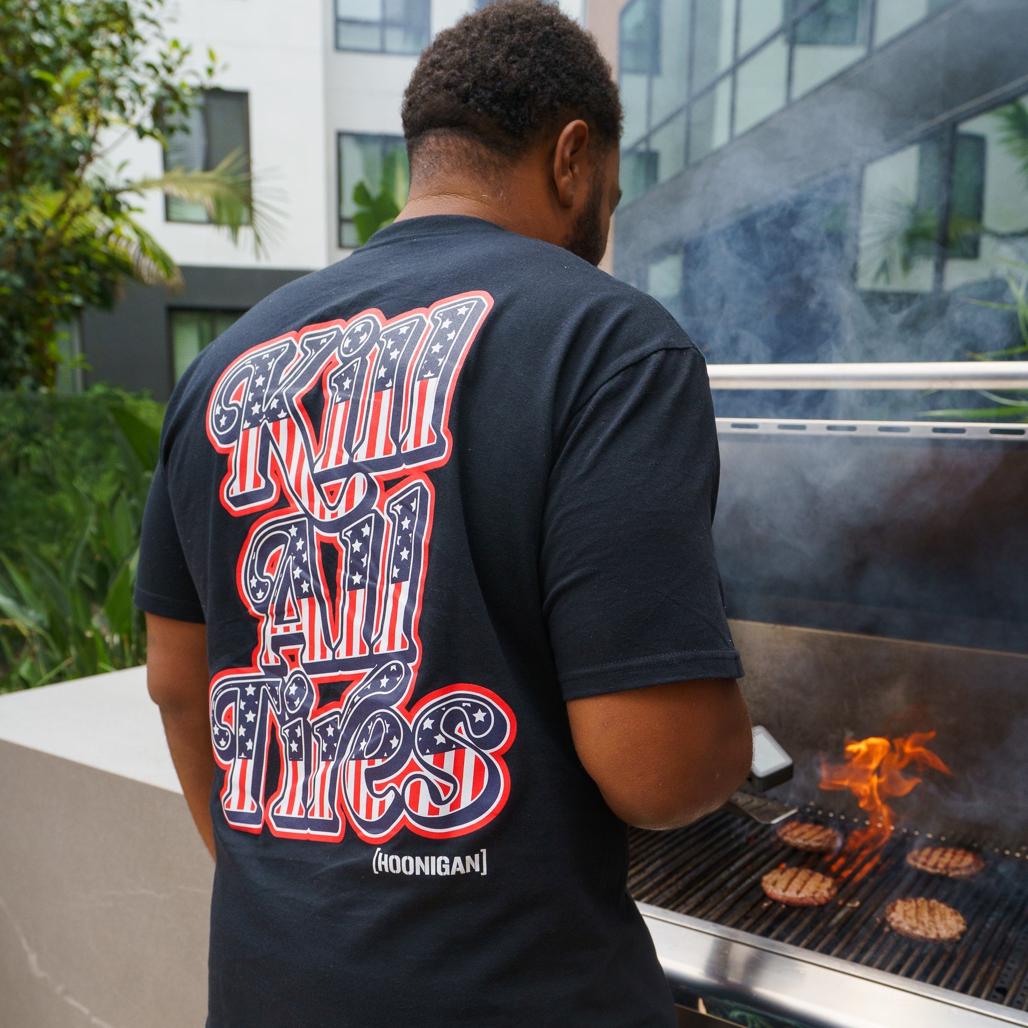 Back view of a man wearing a black Hoonigan Kill All Tires USA short sleeve T-shirt with bold patriotic graphic text, grilling burgers outdoors.