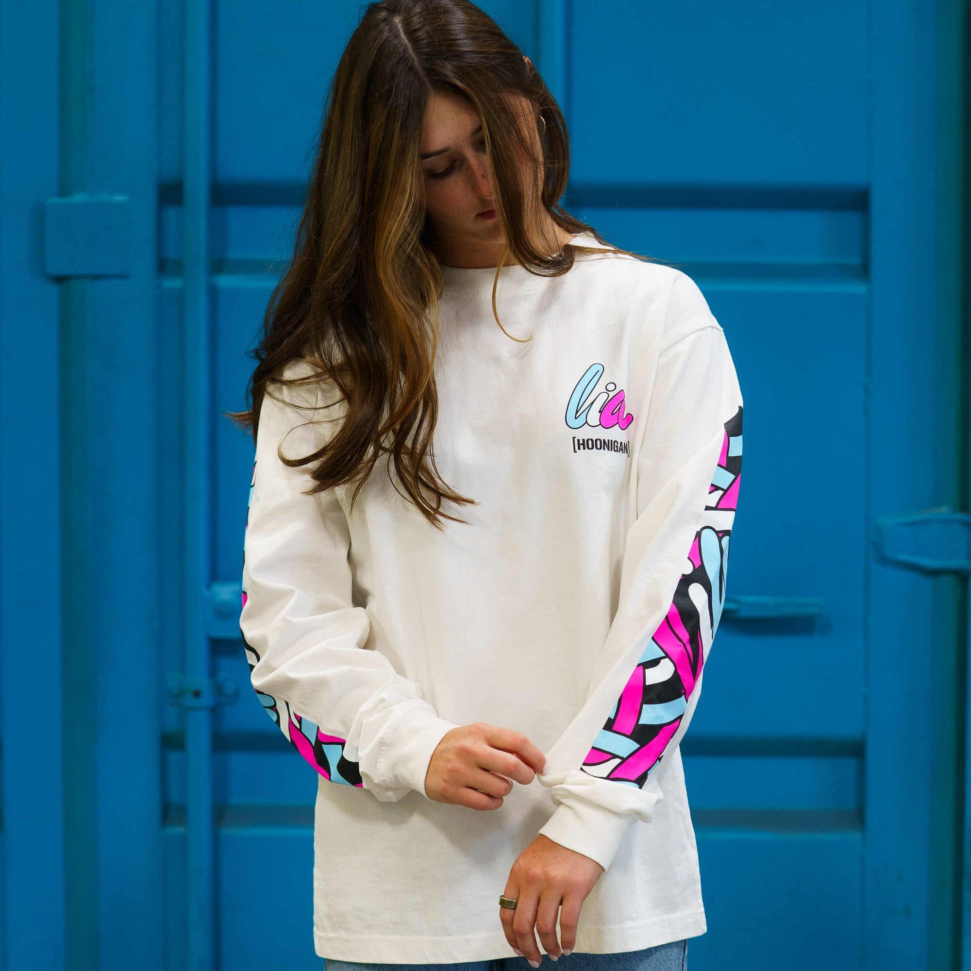 Model wearing Hoonigan x Lia Block SCRIPT Long Sleeve T-Shirt featuring vibrant graphics on sleeves against a blue background.