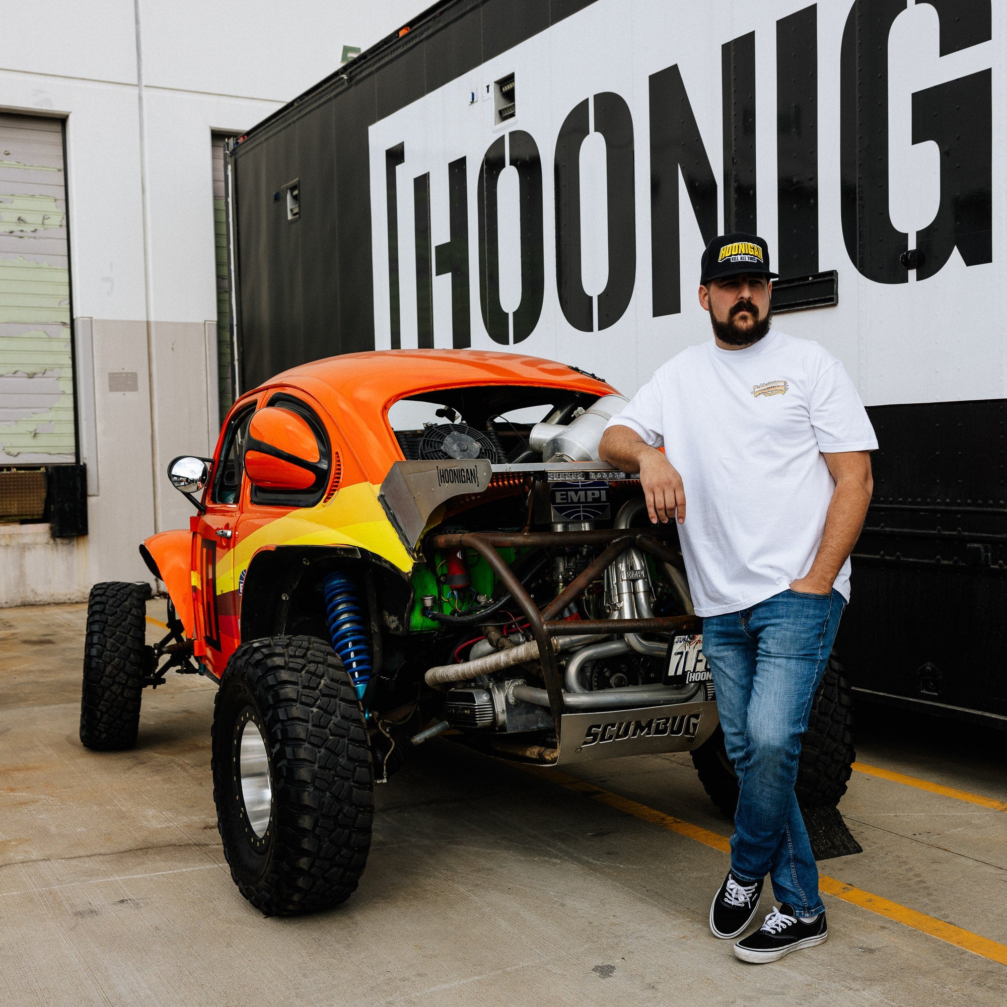 Hoonigan SCUMBUG Short Sleeve T-Shirt worn by a man leaning against a modified off-road vehicle in a garage setting.