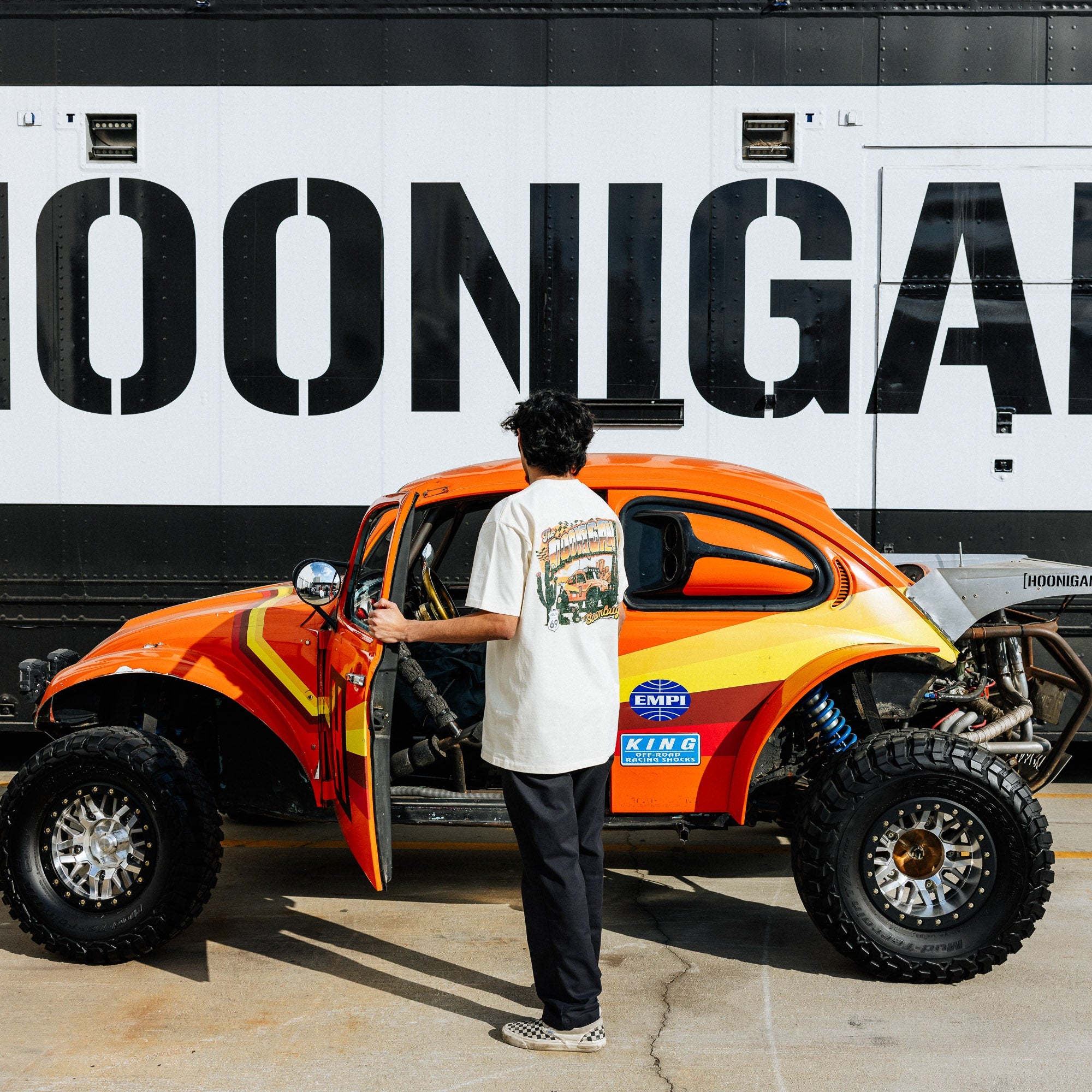 A person stands beside an orange and yellow modified Volkswagen Beetle, wearing a Hoonigan SCUMBUG T-shirt.
